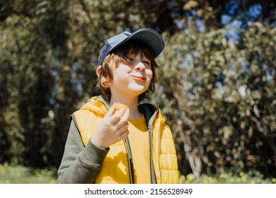 Smiling Cheerful Child Kid In Yellow Vest And Green Hoodie Eats Crisp Snacks Outdoors In Public Park. Schoolboy Boy Enjoying Consumes Chews Junk Food Outside With Trees Vegetation On The Background.