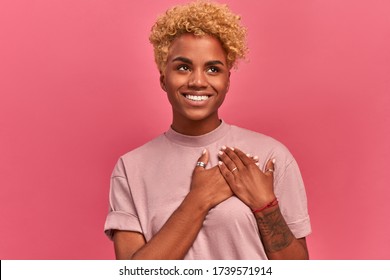 Smiling cheerful dark skinned young female keeps palms on heart, expresses appreciation, pleasure and gratitude, depicts symbol of fight against breast cancer on pink wall. Confession in love concept