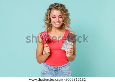Smiling cheerful excited young blonde woman wearing pink basic casual tank top standing hold chocolate bar round lollipop looking camera isolated on blue turquoise colour background studio portrait