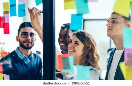 Smiling cheerful female entrepreneur writing funny information on paper stick while enjoying cooperation with male entrepreneur colleagues, multicultural people creating new strategy for proud ceo
