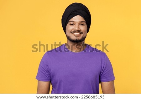 Smiling cheerful fun happy devotee Sikh Indian man ties his traditional turban dastar wear purple t-shirt looking camera isolated on plain yellow background studio portrait. People lifestyle concept