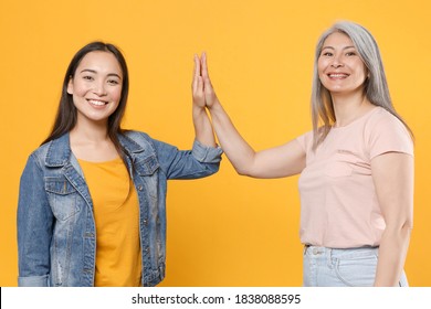 Smiling Cheerful Funny Family Asian Women Gray-haired Mother And Brunette Daughter In Casual Clothes Giving High Five To Each Other Looking Camera Isolated On Yellow Color Background Studio Portrait