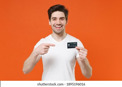 Smiling Cheerful Funny Young Man Wearing Basic Casual Empty Blank White T-shirt Holding In Hands Pointing Index Finger On Credit Bank Card Isolated On Bright Orange Colour Background Studio Portrait