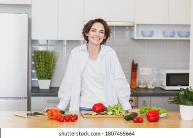 Smiling Cheerful Happy Calm Young Brunette Housewife Woman 20s In White Husband's Shirt And Basic T-shirt Preparing Vegetable Salad, Cooking Food In Light Kitchen At Home Alone.