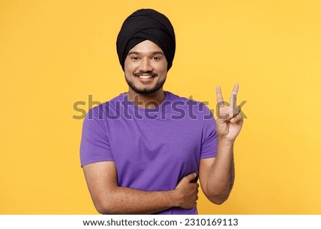 Smiling cheerful happy devotee Sikh Indian man ties his traditional turban dastar wear purple t-shirt showing victory sign isolated on plain yellow background studio portrait. People lifestyle concept