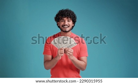 Smiling cheerful happy young bearded Indian man 20s years old wears coral shirt holding showing fan of cash money in dollar banknotes looking camera isolated on plain blue background studio portrait.