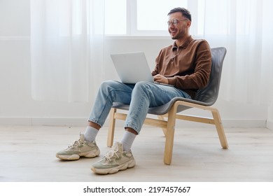 Smiling Cheerful Happy Young Man Guy In Eyewear Looks At Screen Laptop Enjoys Chatting With Friends Doing Cool NFT Project Sitting On Chair At Home. Distance Communication Remote Work New Profession