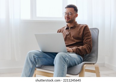 Smiling Cheerful Happy Young Man Guy In Eyewear Looks At Screen Laptop Enjoys Chatting With Friends Doing Cool NFT Project Sitting On Chair At Home. Distance Communication Remote Work New Profession