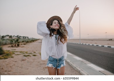 Smiling cheerful long-haired girl with curly hair breathes a full breast and enjoys freedom, standing next to road. Portrait of adorable young woman in white blouse and denim shorts having fun outside - Shutterstock ID 635599844
