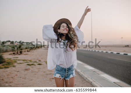 Smiling cheerful long-haired girl with curly hair breathes a full breast and enjoys freedom, standing next to road. Portrait of adorable young woman in white blouse and denim shorts having fun outside