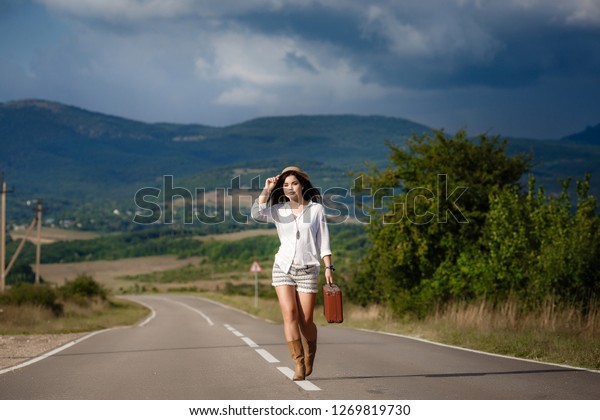 Smiling cheerful long-haired girl with curly hair\
breathes a full breast and enjoys freedom, standing next to road.\
Portrait of adorable young woman in white blouse and denim shorts\
having fun outside