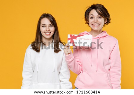 Smiling cheerful pretty two young women friends 20s wearing casual white pink hoodies standing hold gift certificate looking camera isolated on bright yellow color wall background studio portrait