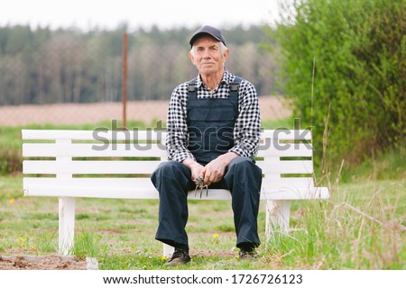Smiling cheerful senior gardener  sitting on bench in garden. Old man in cap and overall resting after work.