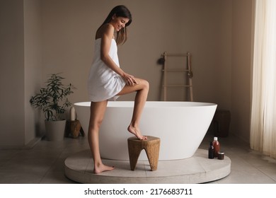 Smiling cheerful woman in towel brushing leg next to big ceramic bathtub, making anti-cellulite or lymphatic drainage massage on hips stepping on wooden stool in luxury bathroom. Body care, spa