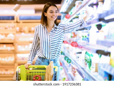 Smiling Cheerful Woman Walking With Shopping Trolley Cart Along The Shelves In Grocery Store. Happy Lady Buying Groceries In Supermarket, Taking Dairy Products Standing Near Fridge Aisle