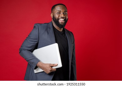 Smiling Cheerful Young African American Business Man 20s Wearing Classic Jacket Suit Standing Hold In Hand Laptop Pc Computer Looking Camera Isolated On Bright Red Color Background Studio Portrait