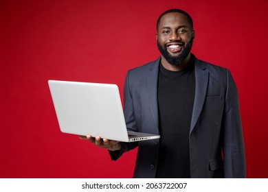 Smiling Cheerful Young African American Business Man 20s Wearing Classic Jacket Suit Standing Working On Laptop Pc Computer Looking Camera Isolated On Bright Red Color Background Studio Portrait