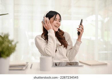 Smiling and cheerful young Asian female in casual clothes enjoying listening to music through headphones in her home working room.