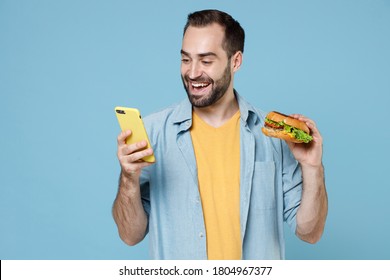 Smiling Cheerful Young Bearded Man 20s In Casual Clothes Holding In Hands American Classic Fast Food Burger And Using Mobile Cell Phone, Typing Sms Isolated On Pastel Blue Background, Studio Portrait.