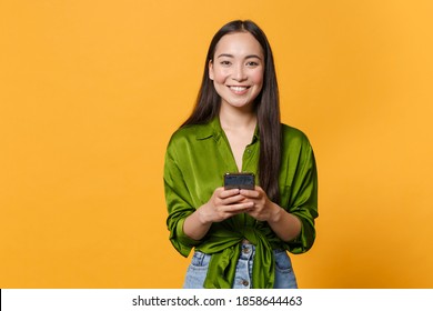 Smiling cheerful young brunette asian woman wearing basic green shirt standing using mobile cell phone typing sms message looking camera isolated on bright yellow colour background, studio portrait Foto Stok