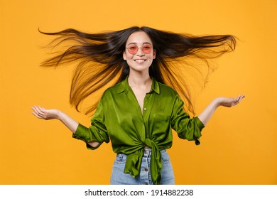 Smiling cheerful young brunette asian woman 20s wearing basic green shirt eyeglasses standing spreading hands throwing fluttering hair isolated on bright yellow colour background, studio portrait
