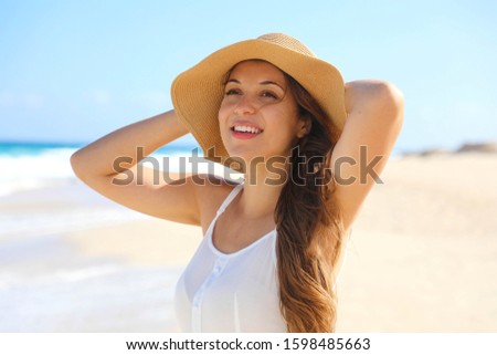 Smiling cheerful young woman with straw hat enjoying wind on the beach