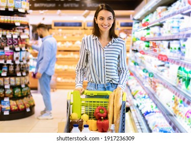 Smiling Cheerful Young Woman Walking With Shopping Trolley Cart Along The Shelves In Grocery Store. Happy Female Customer Buying Groceries In Supermarket, Looking At Fridge With Dairy Products