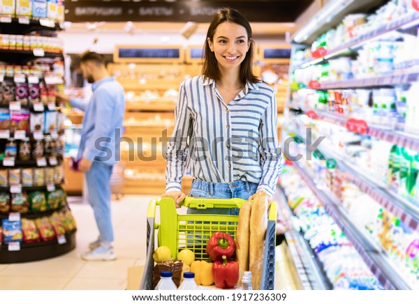 Smiling Cheerful Young Woman Walking With\
Shopping Trolley Cart Along The Shelves In Grocery Store. Happy\
Female Customer Buying Groceries In Supermarket, Looking At Fridge\
With Dairy Products