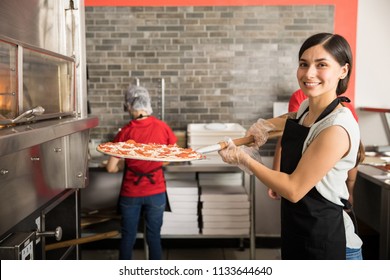 Smiling Chef Wearing Uniform Putting Raw Pizza In Modern Oven For Baking While Looking At Camera And Staff Working In Background In Pizza Shop