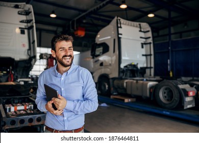 Smiling chief standing in auto park and holding tablet. In background are trucks. Firm for import and export.