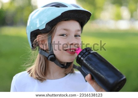 Smiling child girl in a bicycle helmet drinks water from a sports bottle.