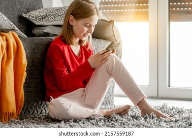 Smiling Child Girl Communicates By Smartphone And Waves Her Hand. Cheerful Kid Spends Time Talking By Video On Celphone Sitting On Floor Near Window.