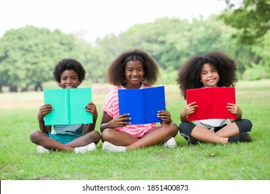 Smiling children holding book outdoor. Happy group of African American children sitting on grass with book and looking to camera outdoor, against green summer garden