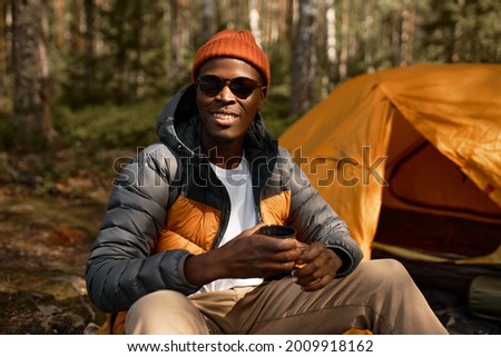 Smiling chilling man camping in mountains. Rest after long walk. Afro american guy enjoy beverage. Virgin nature, contemplating landscape, escape from real world. Travelling concept