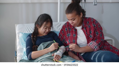 Smiling Chinese Teen Girl Lying In Hospital Bed And Painting Hand Gypsum With Foster Mother. Happy African Adult Woman And Asian Kid Patient Drawing Plaster Case In Hospital Bed