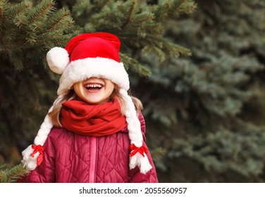 Smiling Christmas Child With A Christmas Hat Pulled Over Her Eyes On Spruce Background