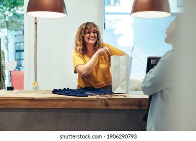 Smiling Clothing Store Owner Packing The Clothes In A Paper Bag Looking At Female Customer And Smiling.