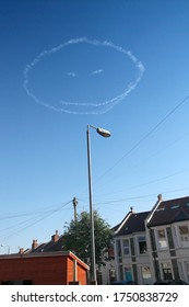A smiling cloud face drawn by a skywriter in an aeroplane looks down from a blue sky onto the houses a suburban neighborhood in the city of Bristol, England.
