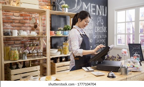 Smiling Coffee Shop Assistant Using Pos Point Of Sale Terminal To Put In Order From Note Paper At Restaurant Register. Waitress Lady Working In Counter In Cafe Store. Young Girl Staff Using Cashbox.