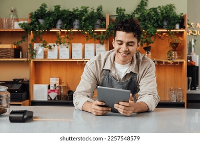 Smiling coffee shop owner at the counter with digital tablet. Male bartender in an apron leaning counter looking at a portable computer. - Powered by Shutterstock