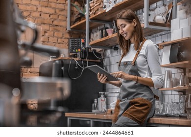 Smiling coffee shop owner woman standing on her workplace near counter using digital tablet. Female barista takes an order from electronic device in a coffee shop. Small business, technology concept. - Powered by Shutterstock