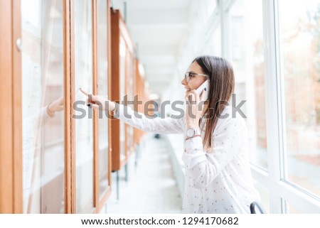 Smiling collage girl with brown hair and eyeglasses using smart phone for calling her colleague to report who passed exams, pointing with finger at noticeboard. University building interior.