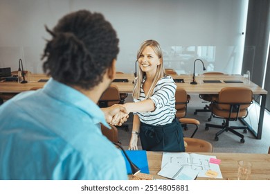 Smiling colleagues greet each other with a handshake in a modern, open-plan office environment, symbolizing teamwork and collaboration. The office features a clean and organized setup.