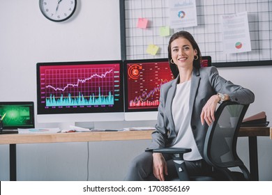 Smiling Computer Systems Analyst Looking At Camera Near Charts On Computer Monitors On Table