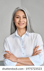 Smiling confident adult 50 years old Asian female professional standing arms crossed looking at camera at gray background. Portrait of sophisticated grey hair woman advertising products and services.