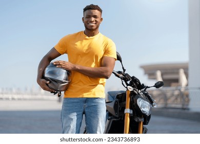 Smiling confident african american man, biker holding helmet and looking at camera near sport motorcycle. Handsome model wearing orange t shirt posing for pictures  - Powered by Shutterstock