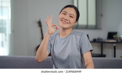 Smiling Confident Asian Woman Showing OK Sign By Hand Sitting On Sofa In Living Room At Home Looking At Camera.