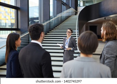 Smiling Confident Business Forum Guide With Badge On Neck Standing On Stairs And Holding Clipboards With Files While Giving Tour To Participants