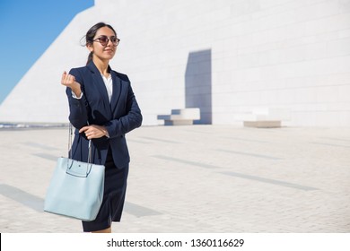 Smiling Confident Business Lady On Her Way To Office. Elegant Young Woman In Formal Suit And Sunglasses With Blue Stylish Bag Walking Outdoors Through Square. Fashion Concept