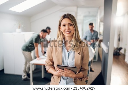 Smiling confident business leader looking at camera and standing in an office at team meeting. Portrait of confident businesswoman with colleagues in boardroom. Posing while holding digital tablet.